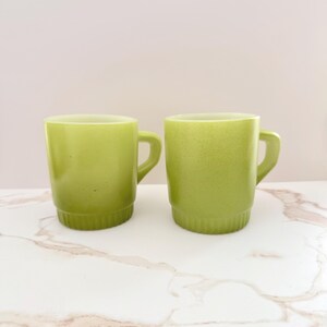 May include: Two green ceramic mugs with handles. The mugs have a textured surface and a stacked base design. The mugs are set on a white marble surface with a light pink background.