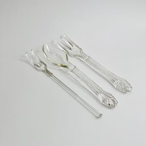 May include: A set of three clear glass utensils consisting of a long, slender two-pronged fork, a central spoon with a smooth oval bowl, and a wider two-pronged fork. Each piece features a textured, leaf-like pattern at the base of the handle.