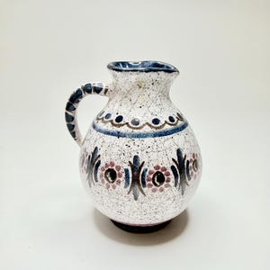 May include: A ceramic pitcher with a rounded body and a single handle, featuring a white base with a dark crackle glaze. The surface is decorated with hand-painted blue and mauve floral motifs, dots, and lines, with a dark blue rim and handle.