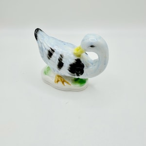 May include: A ceramic duck figurine featuring a glossy white body with light blue speckles and prominent black patches on its wings. The duck has a yellow beak, black eyes, and orange feet, and it is positioned on a small, oval green base.