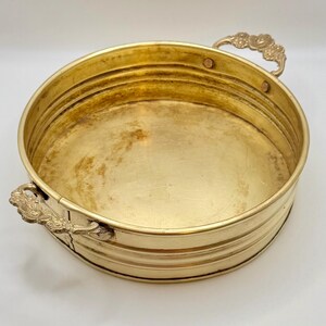 May include: A round, gold-colored metal tray with two ornate handles. The tray has a shallow, flat base and a series of horizontal ridges around the sides. The handles are decorative, with a floral design. The tray is likely used for serving or display.