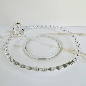 May include: A clear glass serving plate with a beaded rim and a central raised area. The plate has a decorative handle and is designed for serving food or displaying items. The plate is made of clear glass.