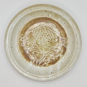 May include: A round, speckled ceramic plate with a raised fish design in the center. The fish is brown, with a textured scale pattern. The plate's rim is a lighter beige color.
