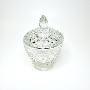 May include: A clear glass candy dish with a matching lid, featuring a textured diamond pattern across the exterior surface. The lid is topped with a pointed finial, and the entire piece rests on a circular base, showcasing a reflective finish.