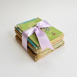 May include: A stack of vintage children's books tied together with a light purple satin ribbon. The top book features a green cover with the text Little Billy Duck and Tiny Elf Library printed in white and red, showing signs of wear on the edges.