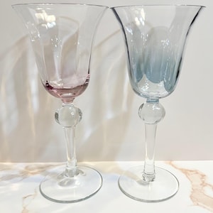 May include: Two elegant wine glasses with flared rims. One glass has a pink hue at the base, while the other has a blue tint. Both glasses have a clear, spherical detail on the stem and sit on a round base. The glasses are made of clear glass.