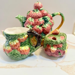 May include: A ceramic tea set featuring a teapot, creamer, and sugar bowl. The teapot is adorned with a cluster of red strawberries, green leaves, and a yellow handle. The creamer and sugar bowl are decorated with strawberries and green leaves.