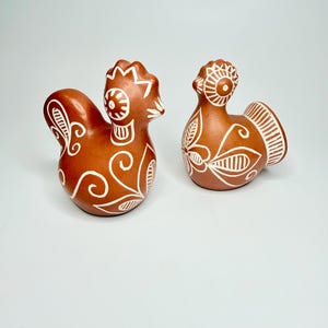 May include: Two terracotta-colored ceramic bird figurines featuring white hand-painted floral and geometric patterns. The figures have rounded bodies, detailed crests, and stylized tail feathers, showcasing intricate line work across their surfaces.