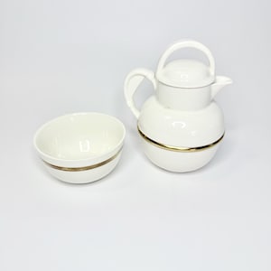 May include: A white ceramic teapot with a matching bowl. The teapot features a rounded body, a curved handle, a lid, and a gold-toned band around the center. The bowl is circular with a matching gold-toned band encircling its exterior surface.