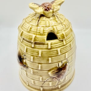 May include: A ceramic honey pot shaped like a traditional straw skep beehive with a glossy light green glaze and a crackle finish. The lid features a sculpted bee handle, and the body is decorated with two brown painted bees in relief.