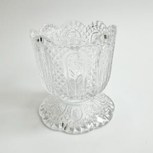 May include: A clear pressed glass bowl with a scalloped rim and a pedestal base. The exterior features a repeating pattern of vertical panels containing embossed flower motifs, separated by sections of a diamond-shaped crosshatch texture.
