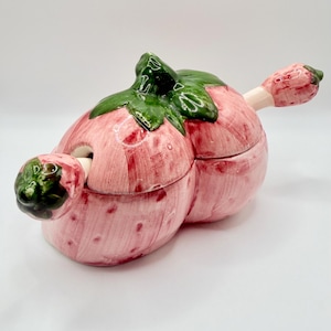 May include: A ceramic lidded jar shaped like a strawberry with a glossy pink finish, green leaves on the lid, and a matching strawberry-shaped spoon handle protruding from the side. The surface features painted red speckles and a smooth texture.
