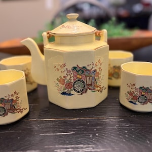 May include: A cream-colored ceramic tea set consisting of a hexagonal teapot with a lid and three matching hexagonal cups. Each piece features a colorful graphic of a traditional cart surrounded by floral patterns and gold-toned trim accents.