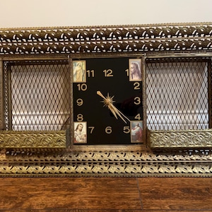 May include: Vintage rectangular wall clock with a gold-toned filigree frame. The clock face is black with gold hands and numerals. Decorative panels with religious imagery flank the clock face. The clock has a decorative gold-toned metal mesh.