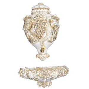 May include: A white and gold decorative wall-mounted water fountain with a grape and leaf design. The fountain has a spigot and a matching basin below. The design is ornate and classical.
