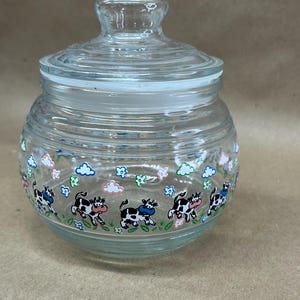May include: A clear glass jar with a lid. The jar is decorated with a repeating pattern of black and white cows, blue clouds, and green flowers. The jar is round with a clear glass lid.