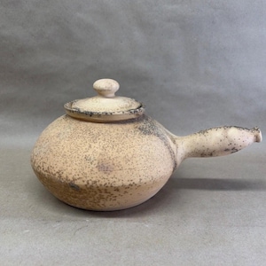 May include: A side-handled ceramic teapot with a rounded body and a matching lid. The piece features a light beige base color with a speckled brown texture throughout, including a darker concentration of spots near the handle and base.