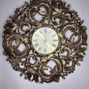 May include: A round wall clock featuring a metallic bronze frame with intricate, swirling scrollwork and floral motifs. The clock face is light-colored with black Roman numerals and thin gold-toned hands. The brand name Elgin is printed in the center.