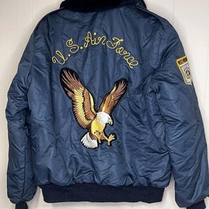 May include: Navy blue U.S. Air Force bomber jacket with a black fur collar and a large embroidered eagle on the back. The words "U.S. Air Force" are embroidered in gold script above the eagle. A patch is on the right sleeve.