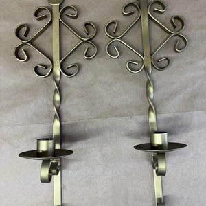 May include: A pair of gold-colored metal wall sconces featuring a vertical bar with a twisted center section, a circular candle holder plate, and an ornate scrollwork top with a pointed finial. Each sconce has a small hole at the top for mounting.