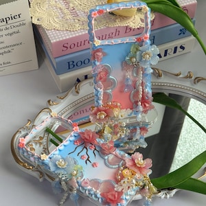 Könnte beinhalten: Eine dekorative Handyhülle mit blau-rosa Farbverlauf, verziert mit 3D-Blumenverzierungen in Rosa-, Blau- und Weißtönen. Die Hülle hat einen Griff und baumelnde Charms, was eine skurrile Ästhetik erzeugt.