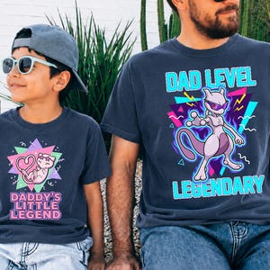 May include: Matching navy blue t-shirts with graphic designs. One shirt features a purple cartoon character with the text "DAD LEVEL LEGENDARY". The other shirt has a cartoon cat with the text "DADDY'S LITTLE LEGEND".