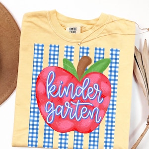 May include: A pale yellow t-shirt featuring a graphic of a red apple with a brown stem and green leaves. The apple is centered over a blue and white gingham striped background. The word kindergarten is written in blue cursive script across the apple.