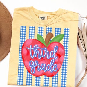 May include: A pale yellow t-shirt featuring a graphic of a red apple with green leaves centered over a blue and white gingham striped background. The text "third grade" is written in a blue cursive font across the center of the apple.