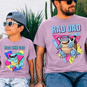 May include: Two matching lilac t-shirts with the phrases "RAD DAD" and "RAD LIKE DAD" in blue and pink. Each shirt features a cartoon turtle wearing sunglasses. The shirts are suitable for a father and child.
