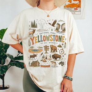 May include: Cream-colored t-shirt featuring a colorful graphic of Yellowstone National Park. The design includes illustrations of wildlife, landmarks, and the park's name. The text "NATIONAL PARK YELLOWSTONE EST. 1872 WYOMING" is also included.