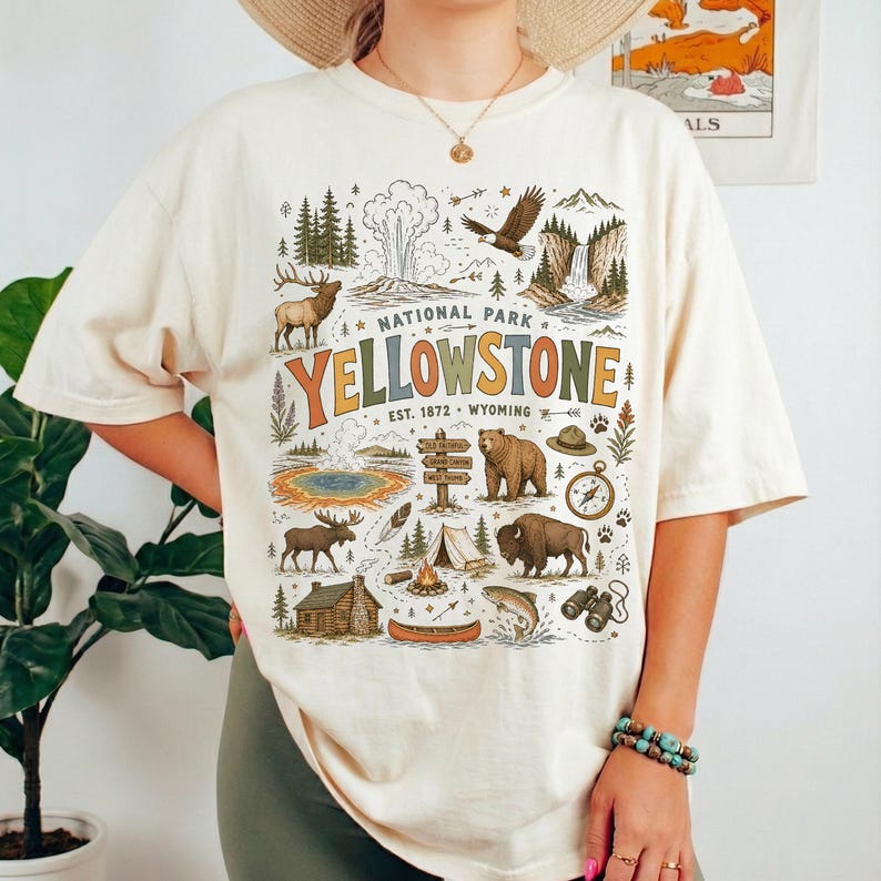 May include: Cream-colored t-shirt featuring a colorful graphic of Yellowstone National Park. The design includes illustrations of wildlife, landmarks, and the park's name. The text "NATIONAL PARK YELLOWSTONE EST. 1872 WYOMING" is also included.