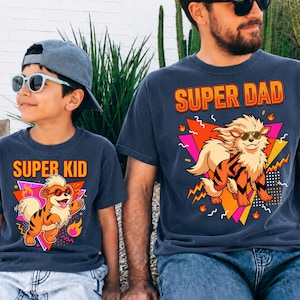 Peut inclure: Deux t-shirts bleu marine avec des graphiques assortis. L'un dit "SUPER DAD" et l'autre "SUPER KID". Chaque t-shirt pr&eacute;sente un tigre de dessin anim&eacute; portant des lunettes de soleil, sur un fond g&eacute;om&eacute;trique avec des accents orange, rose et jaune.