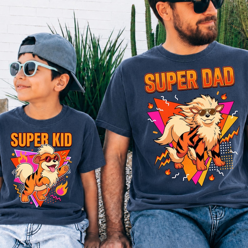 Peut inclure: Deux t-shirts bleu marine avec des graphiques assortis. L'un dit "SUPER DAD" et l'autre "SUPER KID". Chaque t-shirt pr&eacute;sente un tigre de dessin anim&eacute; portant des lunettes de soleil, sur un fond g&eacute;om&eacute;trique avec des accents orange, rose et jaune.