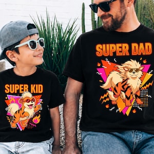 Peut inclure: T-shirts noirs avec des graphiques assortis. L'un des t-shirts affiche "SUPER PAPA" et l'autre "SUPER ENFANT". Chaque t-shirt pr&eacute;sente un chien de dessin anim&eacute; portant des lunettes de soleil, sur un fond g&eacute;om&eacute;trique avec des flammes.