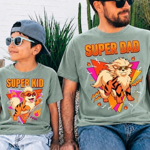Peut inclure: Deux t-shirts vert sauge assortis. L'un dit "SUPER KID" et l'autre "SUPER DAD" en texte orange. Chaque chemise pr&eacute;sente un chien de dessin anim&eacute; portant des lunettes de soleil, sur un fond g&eacute;om&eacute;trique color&eacute;.