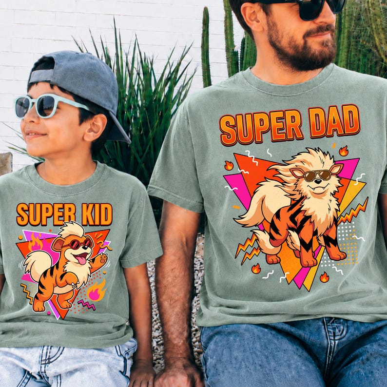 Peut inclure: Deux t-shirts vert sauge assortis. L'un dit "SUPER KID" et l'autre "SUPER DAD" en texte orange. Chaque chemise pr&eacute;sente un chien de dessin anim&eacute; portant des lunettes de soleil, sur un fond g&eacute;om&eacute;trique color&eacute;.