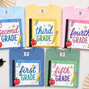 May include: Five t-shirts in light blue, yellow, pink, dark blue, and green. Each features a graphic of a composition notebook with the text second grade, third grade, fourth grade, first grade, or fifth grade, plus a red apple and pencil.