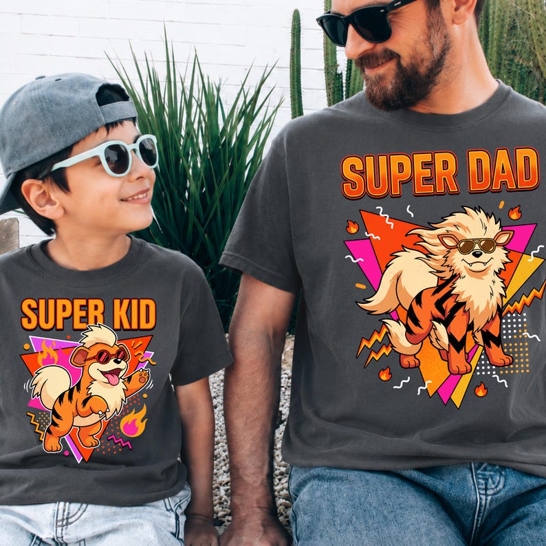 Peut inclure: T-shirts gris anthracite assortis avec les inscriptions "SUPER DAD" et "SUPER KID" en orange, repr&eacute;sentant un chien de dessin anim&eacute; portant des lunettes de soleil. Le chien est orange et noir, avec des flammes et des formes g&eacute;om&eacute;triques.