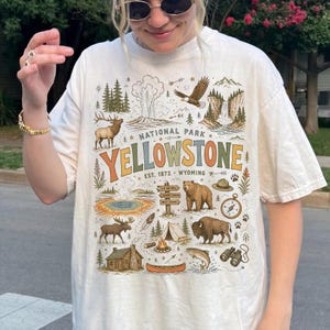 May include: Cream-colored t-shirt featuring a colorful graphic of Yellowstone National Park. The design includes illustrations of wildlife, landmarks, and the text "YELLOWSTONE NATIONAL PARK" with the establishment year 1872.