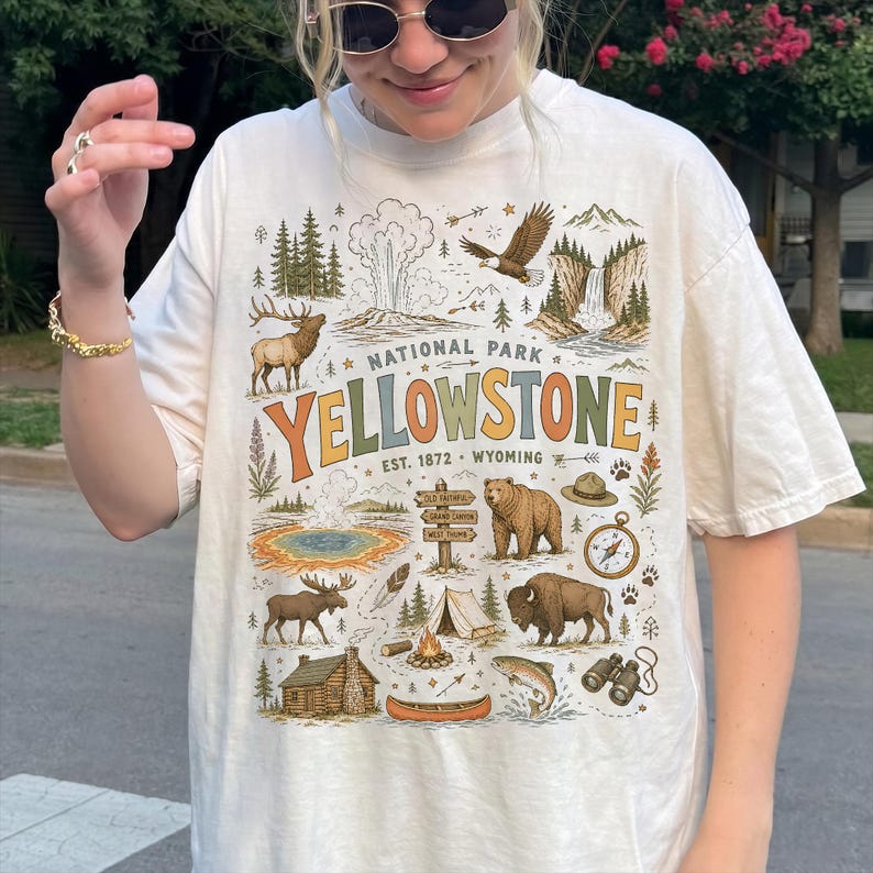 May include: Cream-colored t-shirt featuring a colorful graphic of Yellowstone National Park. The design includes illustrations of wildlife, landmarks, and the text "YELLOWSTONE NATIONAL PARK" with the establishment year 1872.
