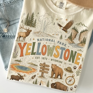 May include: Cream-colored t-shirt with a colorful Yellowstone National Park graphic. The design includes a bison, bear, elk, geyser, and compass. The text reads "National Park Yellowstone, Est. 1872, Wyoming."