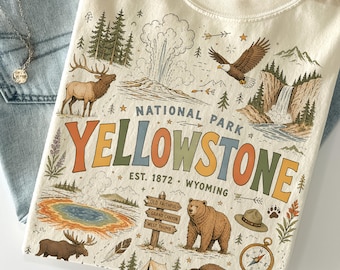 May include: Cream-coloured t-shirt featuring a Yellowstone National Park graphic. The design includes a bison, bear, elk, geyser, and compass. The text reads "National Park Yellowstone, Est. 1872, Wyoming."
