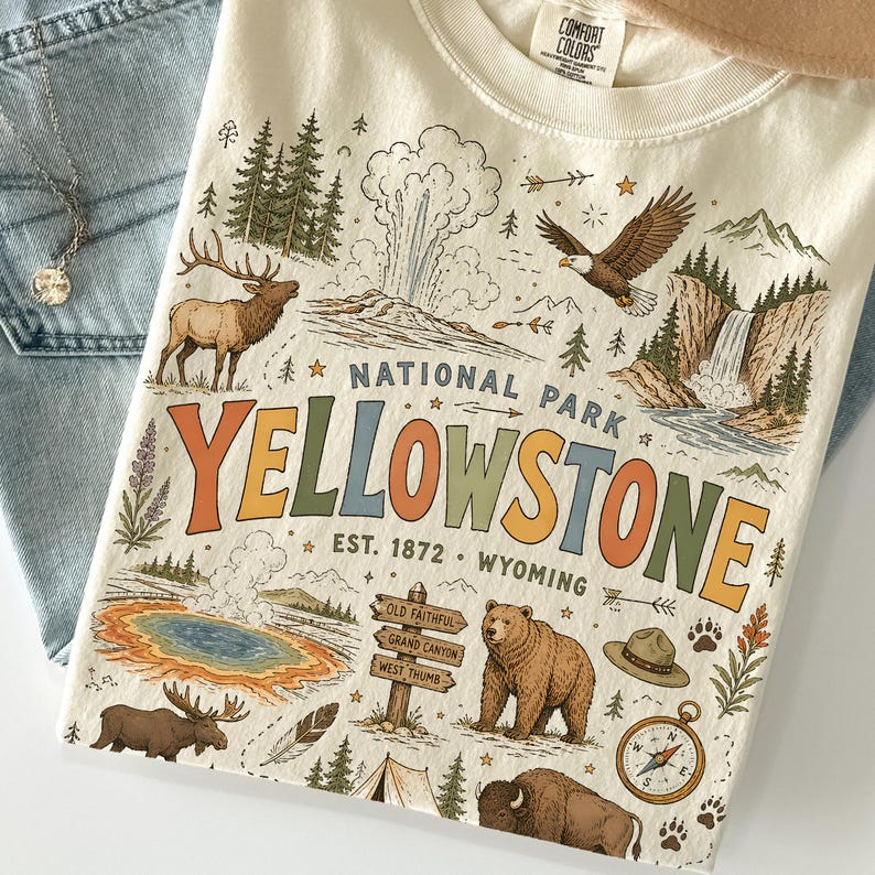 May include: Cream-colored t-shirt with a colorful Yellowstone National Park graphic. The design includes a bison, bear, elk, geyser, and compass. The text reads "National Park Yellowstone, Est. 1872, Wyoming."