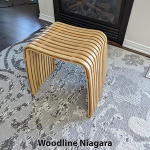 May include: A light brown wooden stool with a curved design. The stool is made of vertical wooden slats and has rounded corners. The text "Woodline Niagara" is at the bottom of the image.
