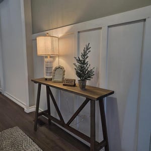 May include: A wooden console table with a lamp, picture frame, and small decorative tree. The lamp has a beige shade and a white base. A small sign reads "Woodline Niagara".
