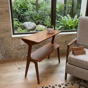 May include: A two-tiered, mid-century modern style side table made of warm-toned wood. The table has a unique, curved leg design and a small, white ceramic bowl on the top shelf. The text "Woodline Niagara" is visible on a small wooden block.