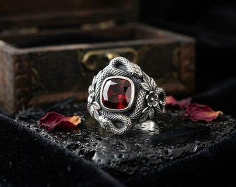 Puede incluir: Un anillo de plata con una gran gema roja cuadrada. El anillo está adornado con intrincados diseños de serpientes y flores. El anillo se muestra sobre una superficie negra texturizada, con una caja de madera borrosa en el fondo.