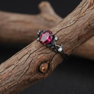 Può includere: Un anello nero con una pietra preziosa ovale rosa intenso come elemento centrale. L'anello presenta un design floreale con piccole pietre bianche ed è posto su uno sfondo di legno. La fascia dell'anello ha una finitura scura e strutturata.