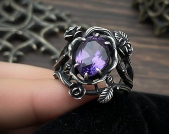 Puede incluir: Anillo plateado con una piedra preciosa ovalada de color púrpura. El anillo presenta un diseño floral, con rosas y hojas. El anillo se muestra sobre un fondo oscuro, resaltando los intrincados detalles de la joya.