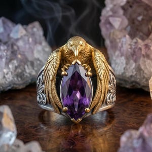Puede incluir: Anillo de plata detallado con detalles dorados, con un dise&ntilde;o de ave que sostiene una gran piedra preciosa p&uacute;rpura con corte marquesa. El anillo se encuentra sobre un fondo de cristales de amatista.